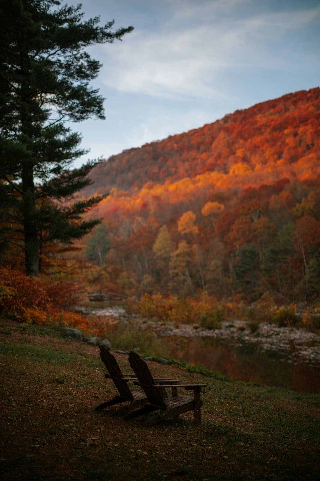 Two Adirondack chairs on The Leeway's creek bank facing a Catskills ridge at peak fall color